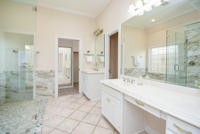 This spacious bathroom features elegant marble accents, a large walk-in shower with glass doors, and dual vanity sinks. The room is bright with natural light from a decorative window, and it has a separate section leading to additional amenities. | Image 29