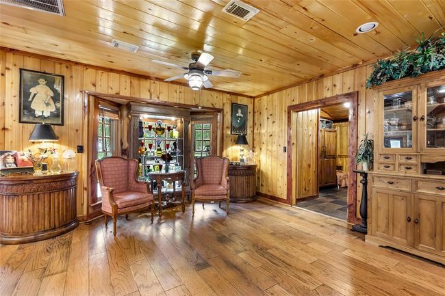 This cozy room features warm wood paneling and flooring, enhancing its rustic charm. It includes elegant seating, decorative shelving by large windows, and ample natural light. A ceiling fan and classic furniture create a welcoming, comfortable space. | Image 22