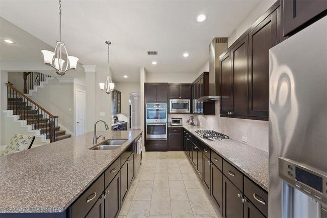 This chef-inspired kitchen boasts rich espresso cabinetry, granite countertops, stainless steel appliances, and a gas cooktop with vent hood. A large island with dual sinks and bar seating anchors the space, while double ovens and a walk-in pantry add convenience. Neutral tile flooring and modern lighting complete the elegant design. | Image 4