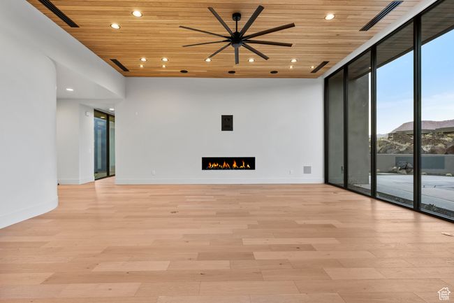 Empty room featuring light wood-style floors, a ceiling fan, floor to ceiling windows, a warm lit fireplace, and wooden ceiling | Image 27
