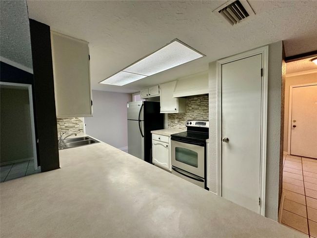 Compact kitchen with modern appliances, stone backsplash, ample countertop space, and overhead lighting. Adjacent to a tiled entryway. | Image 8