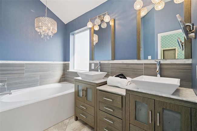 This modern bathroom features a stylish blue accent wall, elegant chandelier lighting, dual vessel sinks with a wood vanity, and a spacious bathtub with sleek gray tile surround. | Image 33
