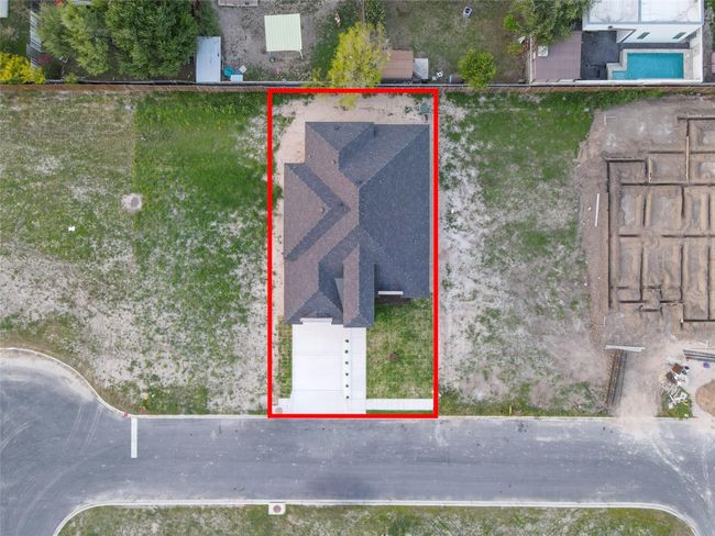 This aerial view shows a newly constructed home with a dark roof, a driveway, and a small front yard. It is situated in a developing residential area with adjacent empty lots and nearby greenery. | Image 4