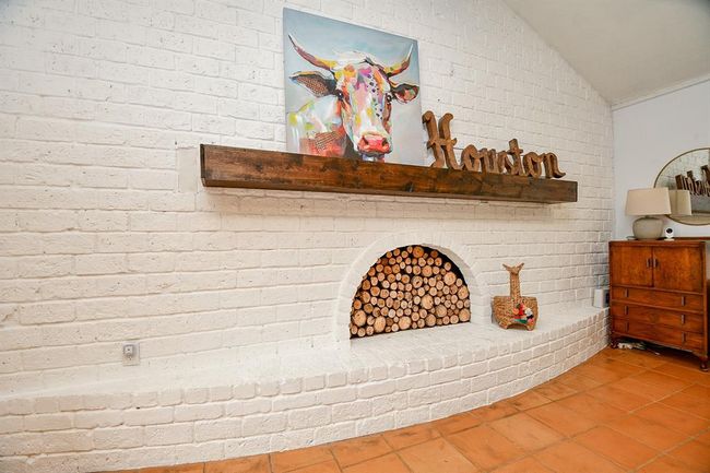 This photo showcases a cozy, rustic-style room with a white brick fireplace filled with logs. The wooden mantel features decorative accents and artwork. Terracotta floor tiles and a vintage wooden dresser add warmth and character to the space. | Image 16