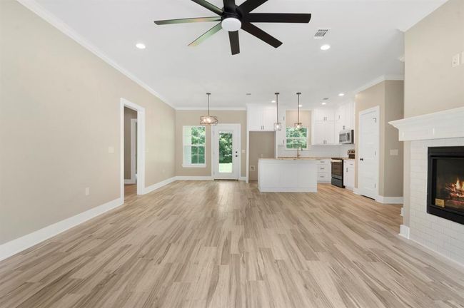 This spacious open-concept living area features modern wood flooring, a cozy fireplace, and a ceiling fan. The kitchen boasts sleek white cabinetry, stainless steel appliances, and an island with pendant lighting. Large windows provide ample natural light, enhancing the room's inviting atmosphere. | Image 6
