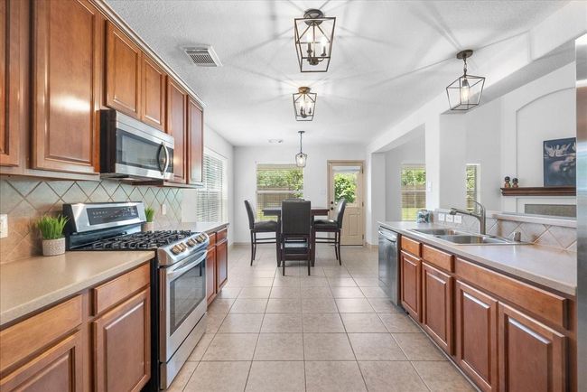 This kitchen features warm wooden cabinetry, stainless steel appliances, and tile flooring. It offers ample counter space and a breakfast area with natural light from large windows. Modern pendant lighting adds a stylish touch. | Image 10