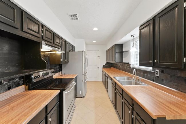 This kitchen features dark cabinetry with wood countertops, stainless steel appliances, and a double sink. It has a galley layout with ample workspace and black tile backsplash, complemented by natural light from a window. | Image 13