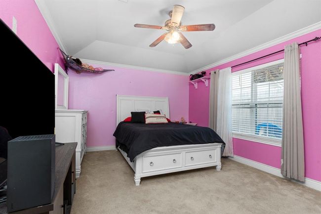 This is a medium-sized bedroom featuring bright pink walls, a white ceiling fan, and a large window with gray curtains. The room is furnished with a white bed frame and matching side drawers, and there is a large TV opposite the bed. The flooring is beige carpet. | Image 25
