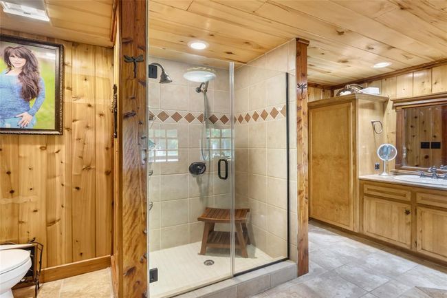 Cozy rustic bathroom featuring wood-paneled walls and ceiling, a glass-enclosed shower with tile accents, and a wooden vanity with a mirror. The space exudes warmth and charm, perfect for a cabin or country-style home. | Image 31