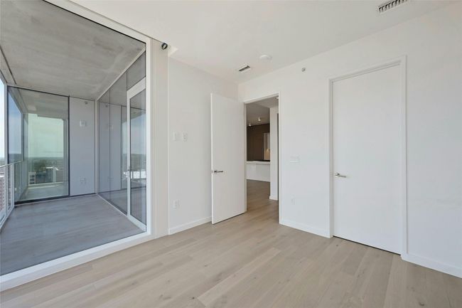 Bright and airy bedroom showcasing expansive floor-to-ceiling windows that frame stunning city views. Light wood flooring and clean, modern finishes create a sleek and tranquil retreat. The room offers easy access to the main living area and includes a spacious closet for added functionality. | Image 12