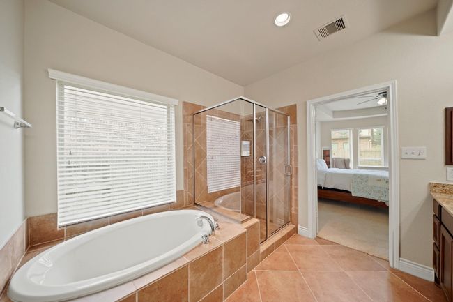 This bathroom features a spacious layout with a large oval bathtub and a glass-enclosed shower. It has tiled flooring and walls, a window for natural light, and an adjoining entrance to a bedroom with carpeted flooring. | Image 23