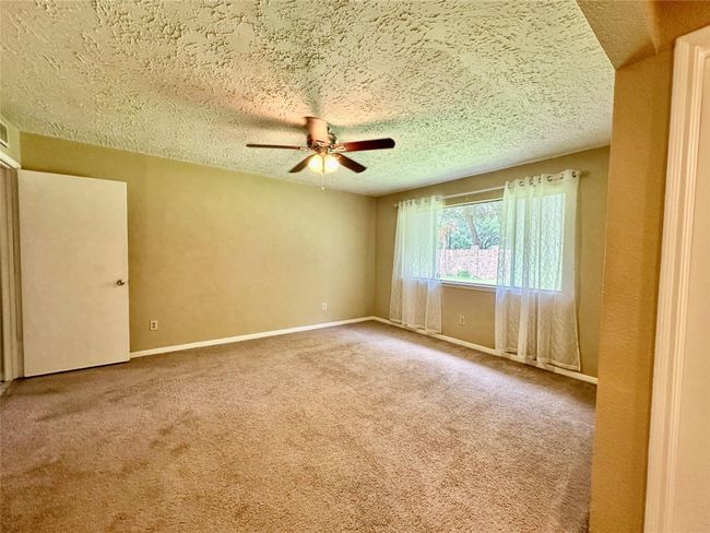 This room is spacious and well-lit, featuring a large window with sheer curtains, a ceiling fan, and neutral carpet and walls, offering a versatile space for any home decor. | Image 9