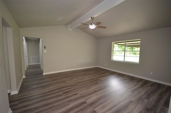 This spacious family room features modern wood-style flooring, neutral walls, and a vaulted ceiling with a fan. A large window provides ample natural light, while the open layout leads to a hallway, offering a versatile living area. | Image 11