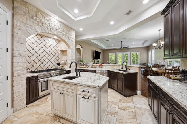 This spacious kitchen features elegant stone accents, a large island with dual sinks, and modern appliances. It opens to a bright living and dining area with hardwood floors, offering a seamless flow for entertaining. | Image 6