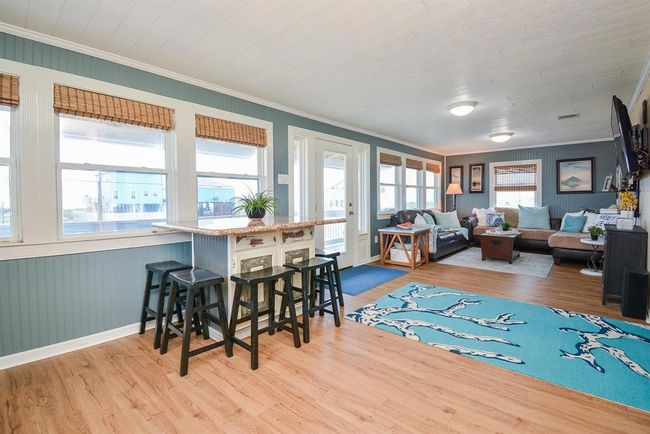 This bright, airy living space features large windows, light wood flooring, and a cozy seating area. The open layout includes a breakfast island with seating, and the room has a relaxed, coastal vibe, ideal for comfortable living and entertaining. | Image 8