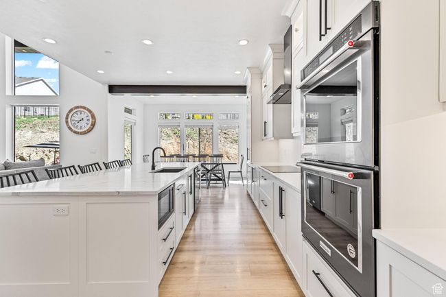 Kitchen featuring light wood-type flooring, a kitchen island with sink, wall chimney exhaust hood, double oven, and a sink | Image 57