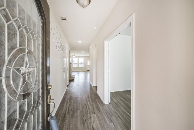 This photo shows a bright entryway with wood flooring and a decorative glass front door. The hallway leads to a spacious living area with large windows and a ceiling fan, providing a welcoming and open atmosphere. | Image 6