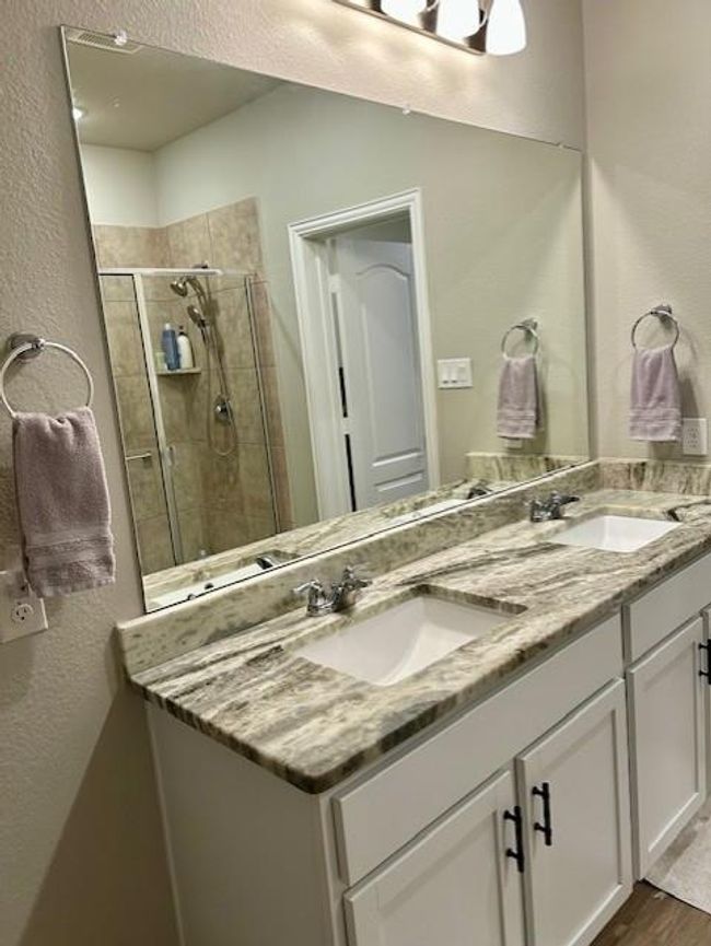 This bathroom features a spacious double vanity with granite countertops and modern fixtures. There’s a large mirror above the sinks, and a glimpse of a tiled shower with glass doors in the background. The neutral color palette adds a clean, contemporary feel. | Image 16