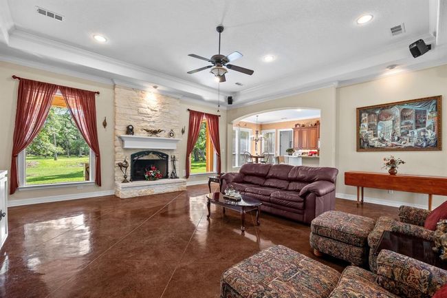 Vaulted tray ceiling, full-height limestone fireplace and recessed lighting frame this inviting gathering space—rich drapes and oversized windows flood the room with natural light and woodland views. | Image 13