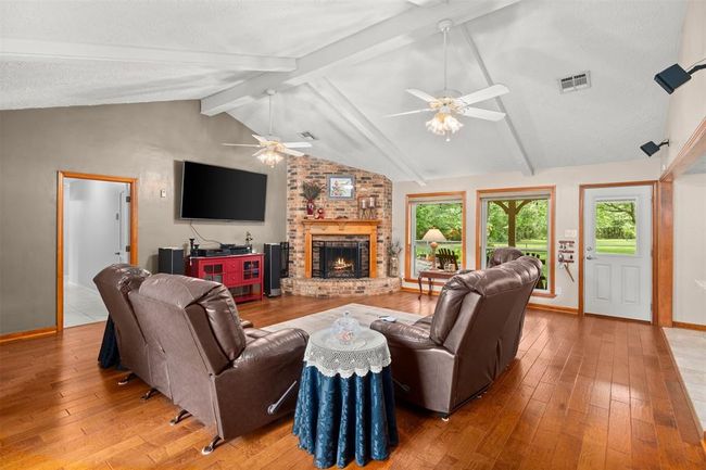 This cozy living room features a charming wood burning fireplace with a rich wooden mantel, complemented by a large window offering scenic views of the lush backyard. The space includes hardwood flooring and is well-lit, providing a warm and inviting atmosphere. | Image 5