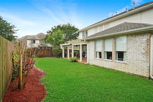 Spacious backyard with lush green lawn and a wooden privacy fence. Features a partially covered patio area, perfect for outdoor relaxation and entertaining. The home exterior is light brick with large windows offering plenty of natural light. | Image 29