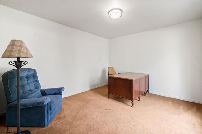 This is a modestly sized, unfurnished room with beige carpeting, white walls, and a simple ceiling light fixture. There's a blue upholstered chair and a floor lamp on one side, and a wooden desk on the other. The space is clean and appears to be in good condition, offering a blank canvas for personalization. | Image 6