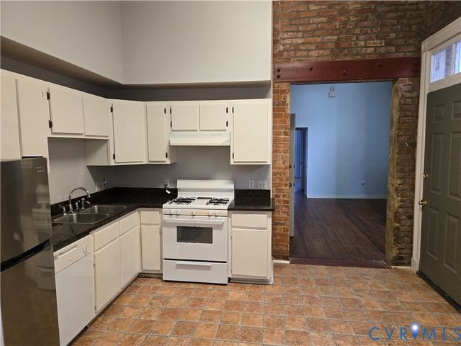 Kitchen with white appliances, dark countertops, white cabinets, under cabinet range hood, and brick wall | Image 9
