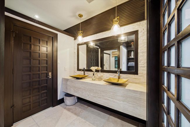 Inside the Zen-inspired air-conditioned pool house, this refined bathroom showcases sleek, spa-like finishes with a floating stone vanity, dual sculptural vessel sinks and striking pendant lighting. A textured stone accent wall adds depth and warmth, while the dark wood trim and custom grid doors maintain a cohesive aesthetic with the home's architectural style. Thoughtfully designed, it offers a luxurious and functional space for post-swim refreshment. | Image 47