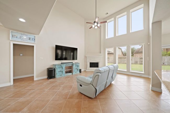 Spacious living room with high ceilings, large windows for natural light, and a central fireplace. The room features tile flooring, and a ceiling fan. The open layout provides a cozy yet airy feel. (TV doesn't convey). | Image 12