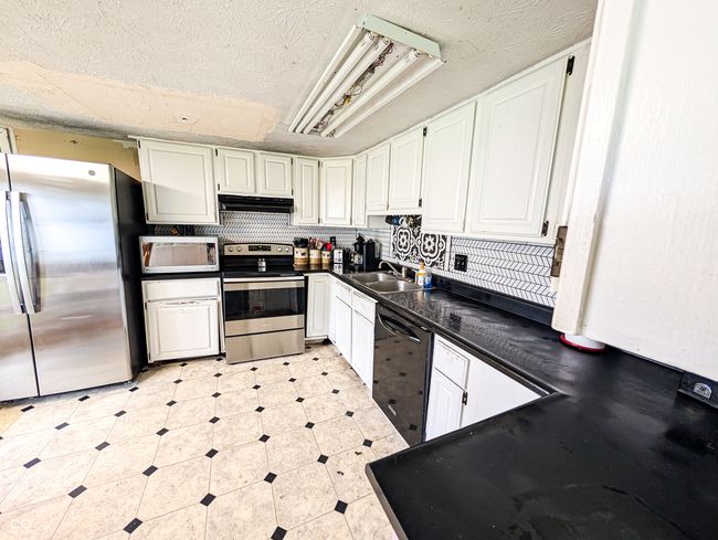 kitchen with stainless steel appliances, under cabinet range hood, a sink, dark countertops, and white cabinetry | Image 4