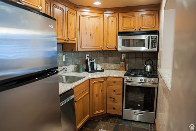 Kitchen with stainless steel appliances, brown cabinetry, backsplash, and light stone counters | Image 11