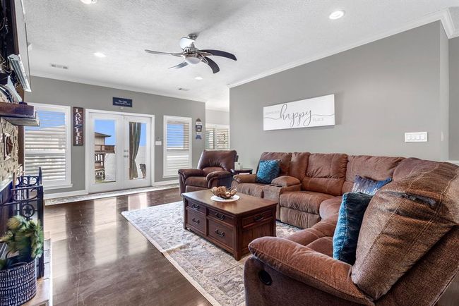 Bright and inviting living room with neutral tones, featuring a comfortable brown sectional sofa, accent chairs, and a stylish ceiling fan. Large windows and glass doors let in ample natural light, highlighting the cozy decor. The room opens to a view of an outdoor space, perfect for relaxation and gatherings. | Image 6