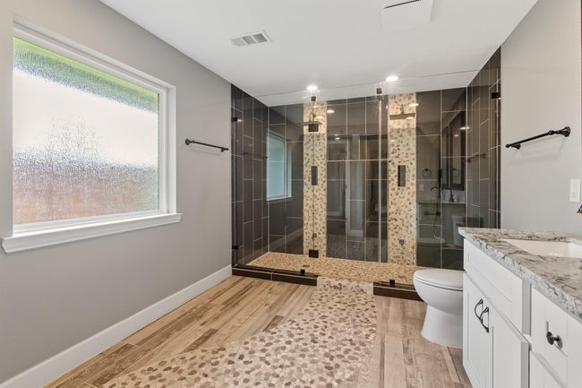 The primary bathroom is sure to give you that spa like feel. It has a large walk in glass shower with double shower heads and controls. The shower has pebbled walkway and floor and large black tile walls. | Image 18