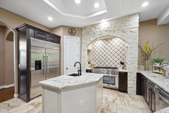 This kitchen features a modern design with stainless steel appliances, a large double-door refrigerator, and a central island with a sink. It has stone accents around the stove, ample counter space, and a warm, inviting color palette. | Image 7