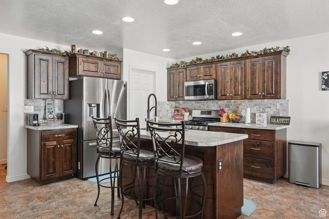 Kitchen featuring tasteful backsplash, light countertops, appliances with stainless steel finishes, a kitchen island with sink, and dark brown cabinetry | Image 17