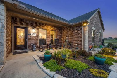 This image shows a charming brick home with an inviting entryway, featuring a decorative door and a cozy porch with seating, overlooking a well-manicured landscape with a view of an open green space. | Image 3