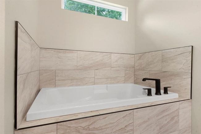 Modern bathroom with a sleek, white tub surrounded by beige marble tiles and featuring stylish black fixtures. A small window above provides natural light. | Image 16