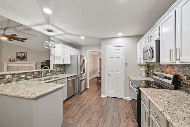 Continue into a stylish & functional kitchen with granite countertops, stainless steel appliances, white cabinetry, and a mosaic tile backsplash. Wood-look tile flooring and modern lighting tie it all together in this open-concept layout. | Image 5