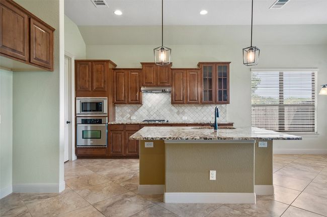 Kitchen with light stone countertops, backsplash, stainless steel appliances, brown cabinetry, and glass insert cabinets | Image 18