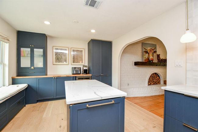 This kitchen features sleek blue cabinetry, a spacious marble-topped island, and light wood floors. A cozy built-in bench sits by the window, and an arched entryway leads to a charming brick nook with a decorative fireplace. | Image 20