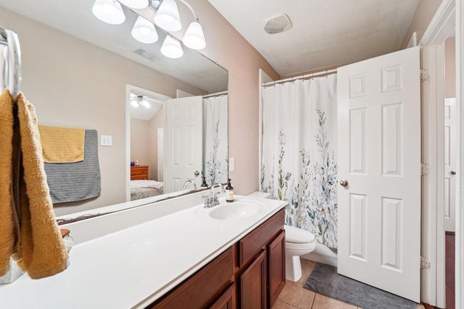 This photo showcases a clean, well-lit bathroom featuring a large mirror, modern light fixtures, and a spacious countertop with wooden cabinetry. The shower area is adorned with a floral-patterned curtain, and there's a visible doorway leading to an adjacent room, providing convenience and accessibility. | Image 19