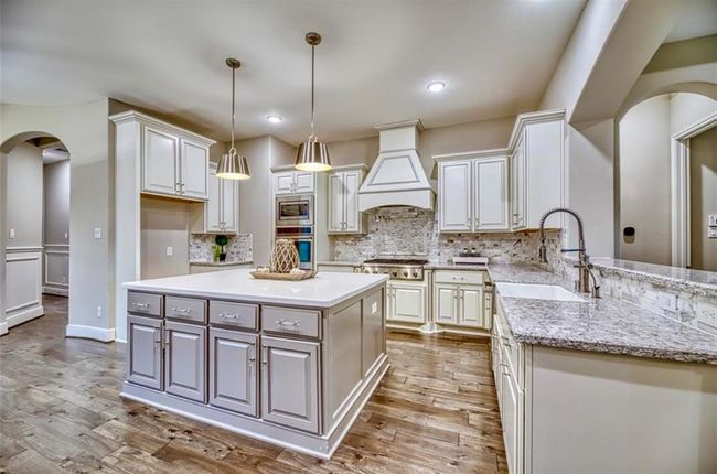 This kitchen boasts a spacious, open design with elegant white cabinetry and granite countertops. It features a large central island, stainless steel appliances, pendant lighting, and a stylish tile backsplash, all set against warm hardwood floors. | Image 23