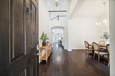 This photo shows an elegant entryway with dark hardwood floors, leading into a bright, spacious dining area with a chic chandelier, followed by a glimpse of a cozy living space. The home features high ceilings and a neutral color palette, promising a modern and airy feel. | Image 3