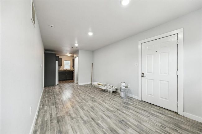 This photo shows a bright, narrow room with modern, laminate flooring and neutral walls. The space leads into a kitchen area with new cabinetry and granite countertops. There's a laundry door on the right, for the laundry room. Overall, it presents a clean and open layout. | Image 4