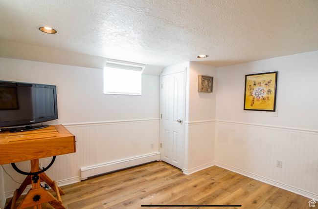 Basement featuring a baseboard heating unit, a textured ceiling, light wood finished floors, a wainscoted wall, and baseboards | Image 12