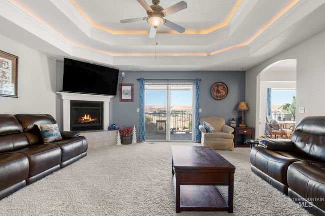 Carpeted living area with a doubletray ceiling, a ceiling fan, ornamental molding, a gas log fireplace, arched walkways & sliding glass door to covered upper deck with beautiful views | Image 8