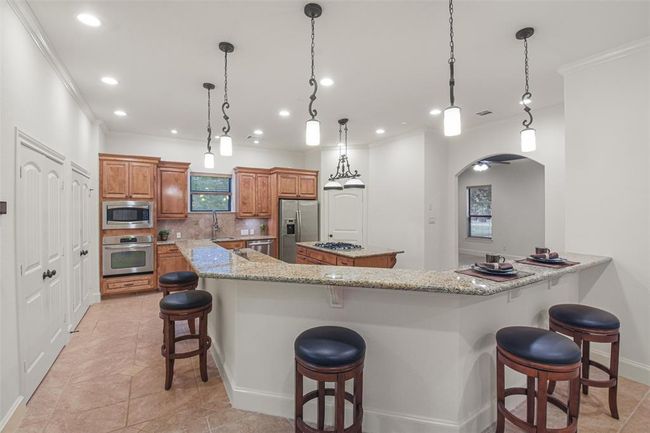 This kitchen features rich wooden cabinetry, granite countertops, modern stainless-steel appliances, a breakfast bar with seating, and elegant pendant lighting, all set on tiled flooring. | Image 18
