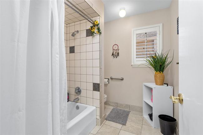 This secondary bathroom includes a tiled shower and bathtub combination, accented with decorative tile inlays. A window with plantation shutters allows for natural light while maintaining privacy. The neutral-toned walls and flooring create a warm and inviting atmosphere. A built-in grab bar adds accessibility, and the layout provides ample space for storage and functionality. | Image 31