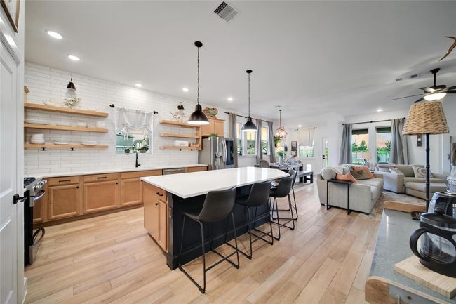 This photo showcases a spacious, open-concept kitchen and living area. The kitchen features a large island with seating, modern pendant lighting, and open shelving against a subway tile backsplash. The living space is bright and inviting, with ample natural light from large windows and a cozy seating area, making it ideal for entertaining and family gatherings. | Image 16
