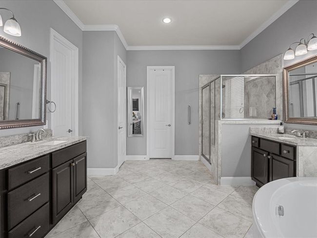 This spacious primary bathroom features dual vanities with elegant granite countertops, a large glass-enclosed shower, and a separate soaking tub. The neutral tile flooring and soft gray walls create a calming atmosphere, complemented by modern lighting and ample storage. | Image 24